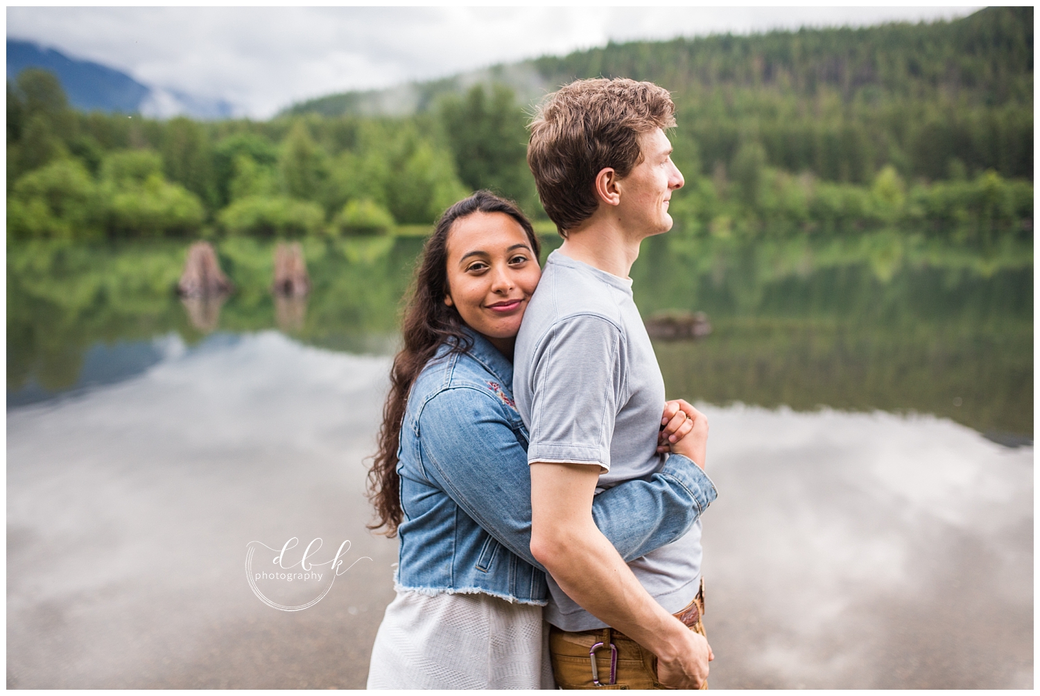 woman hugs her fiance from behind as they stand in front of Rattlesnake Lake in North Bend, Washington