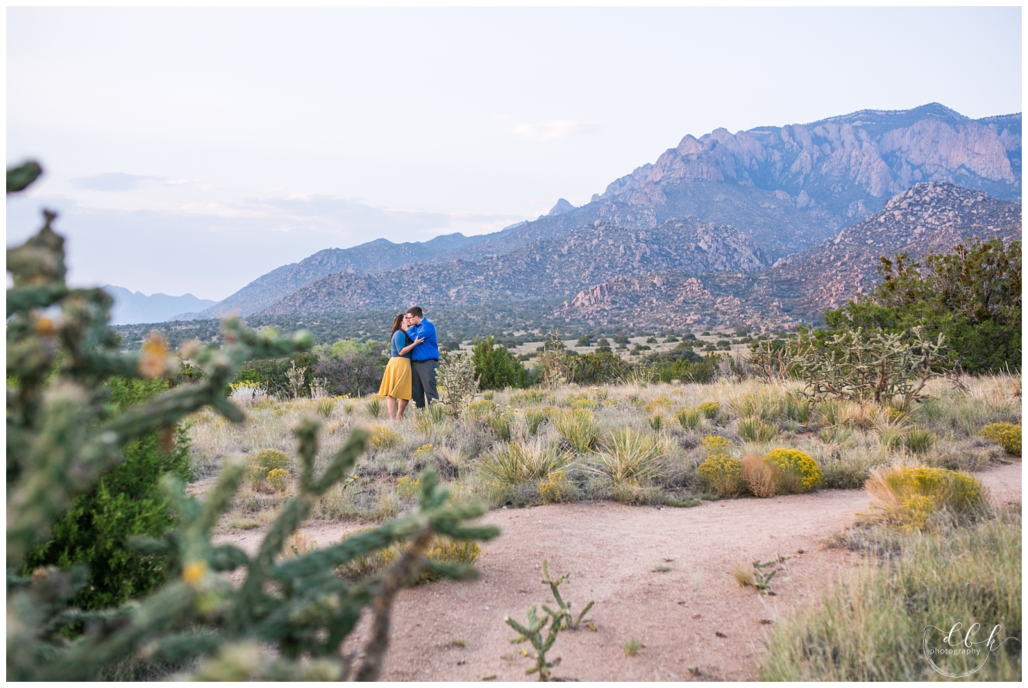 couple wearing blue and yellow kissing for engagement portraits by Sandia mountains in Albuquerque, New Mexico