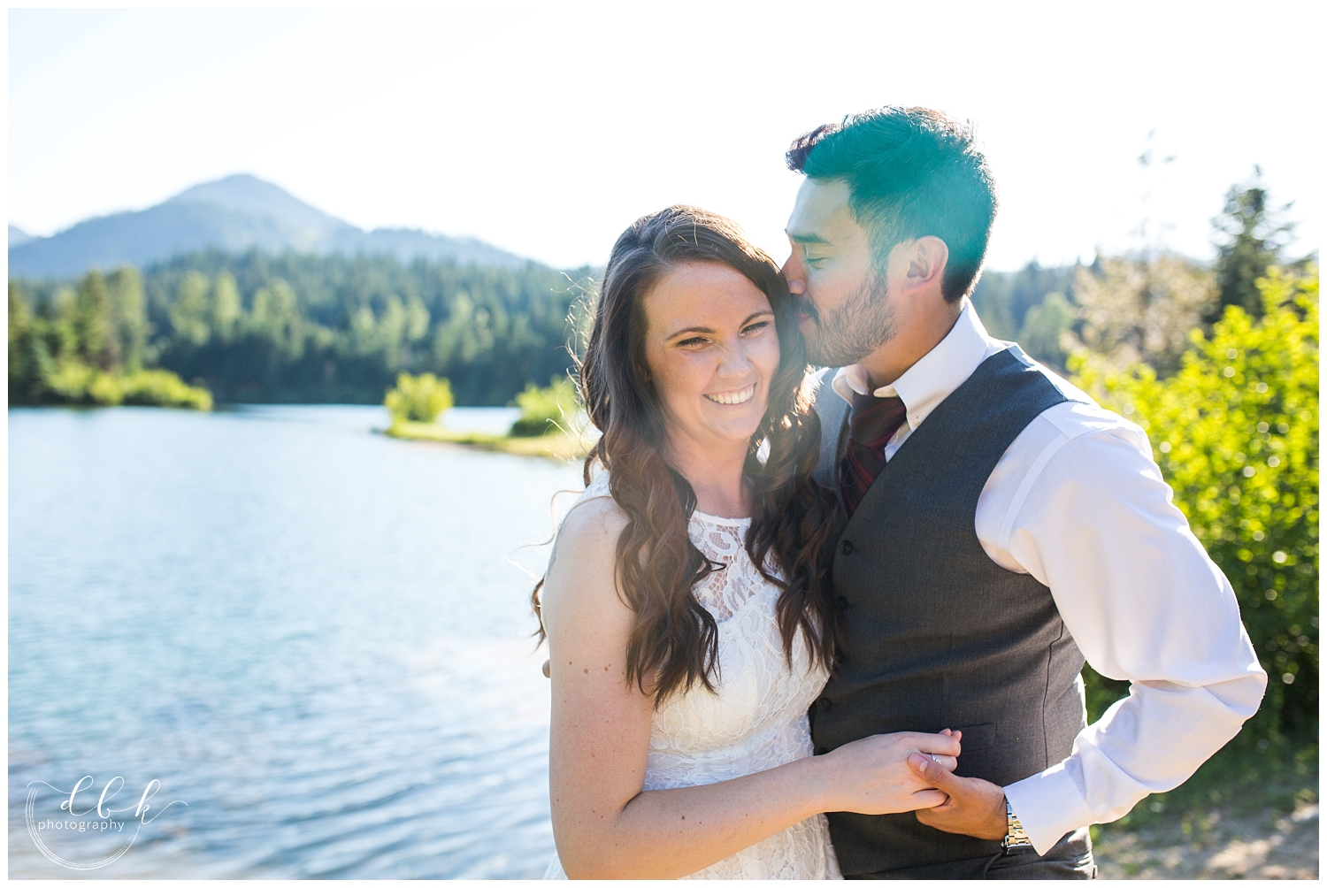 groom in tie and vest kisses bride on the head by the water at Gold Creek Pond