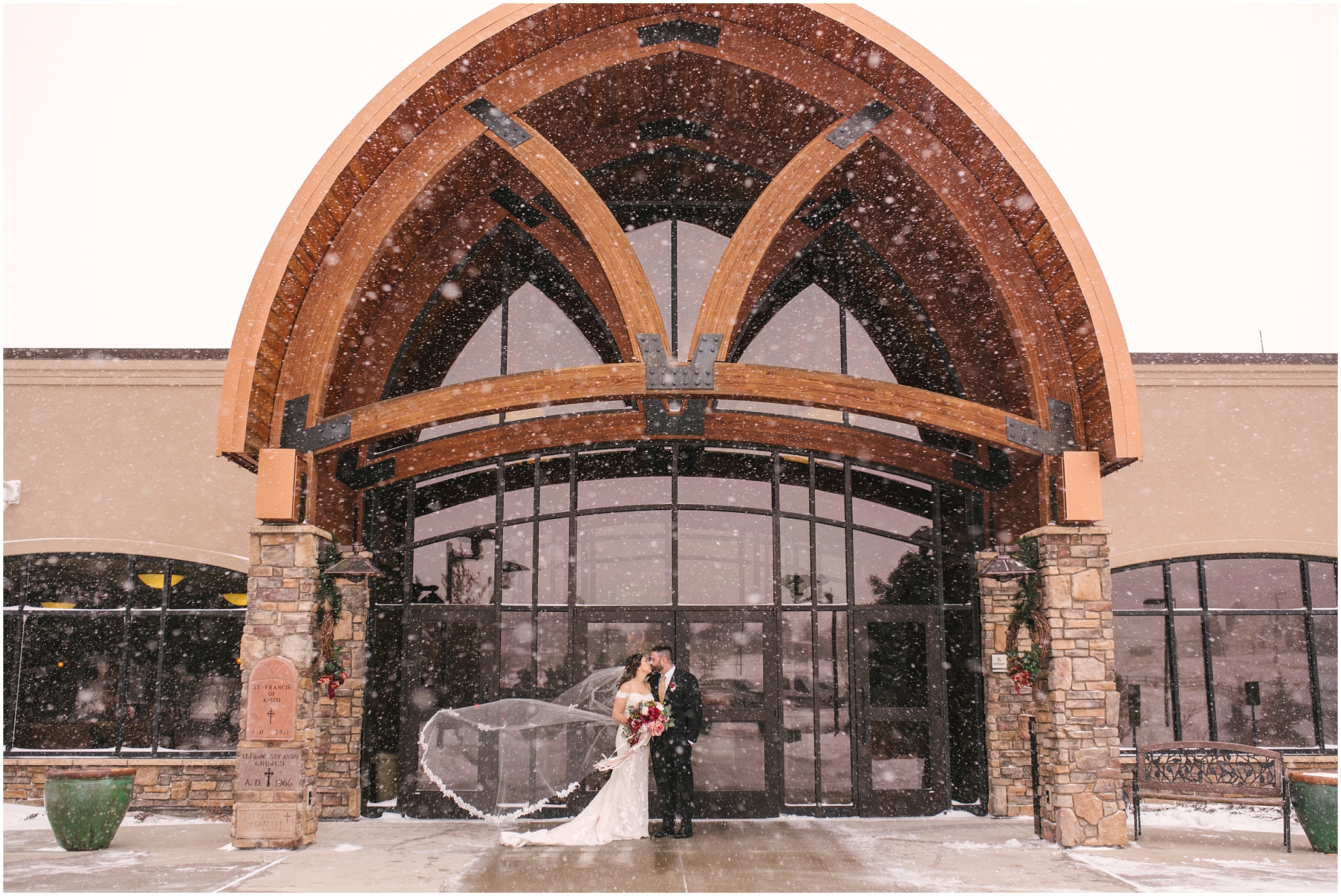 Bride and groom kissing as bride's veil blows behind her in the snow outside St Francis of Assisi Catholic Church
