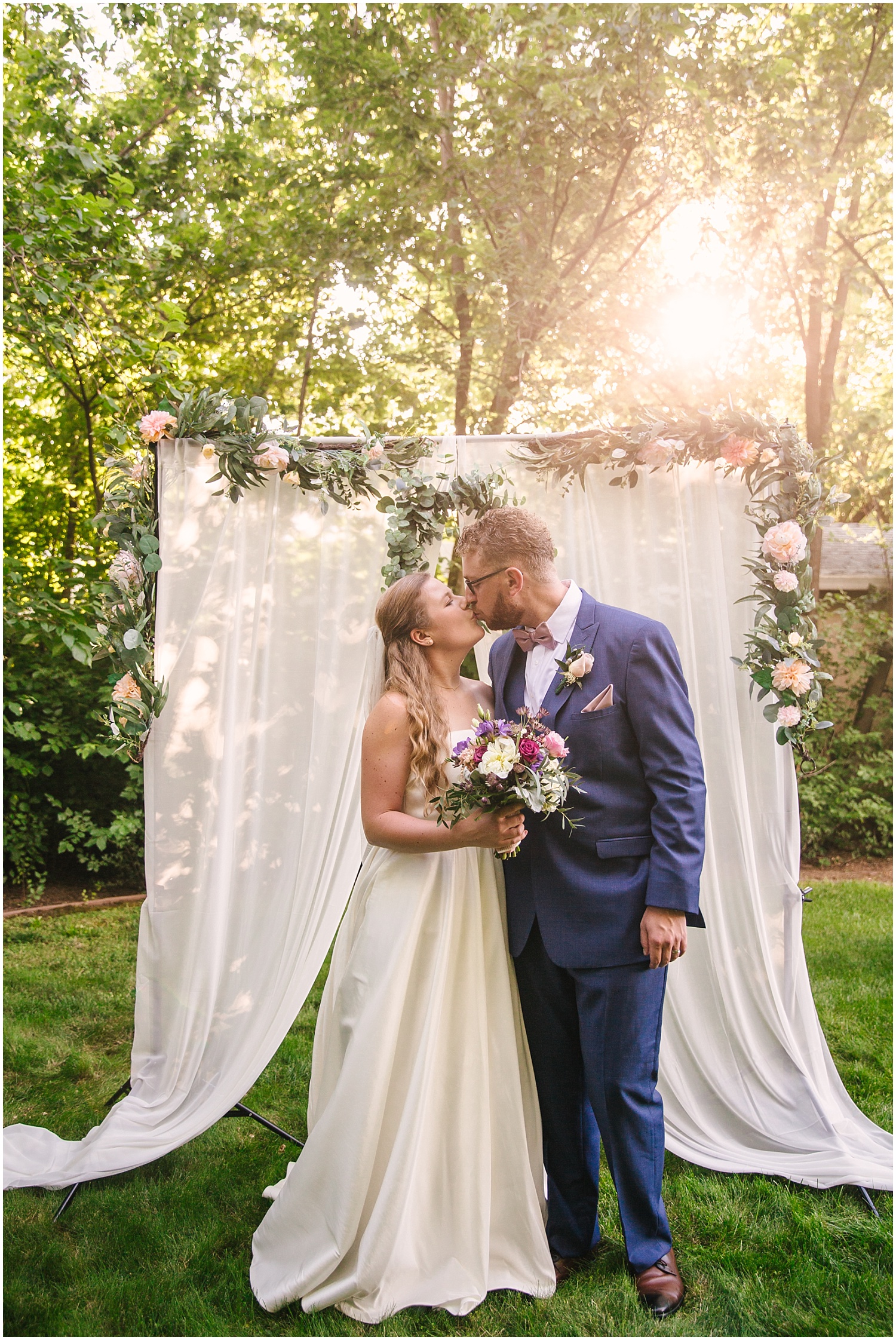 Bride and groom kissing at golden hour surrounded by lush greenery
