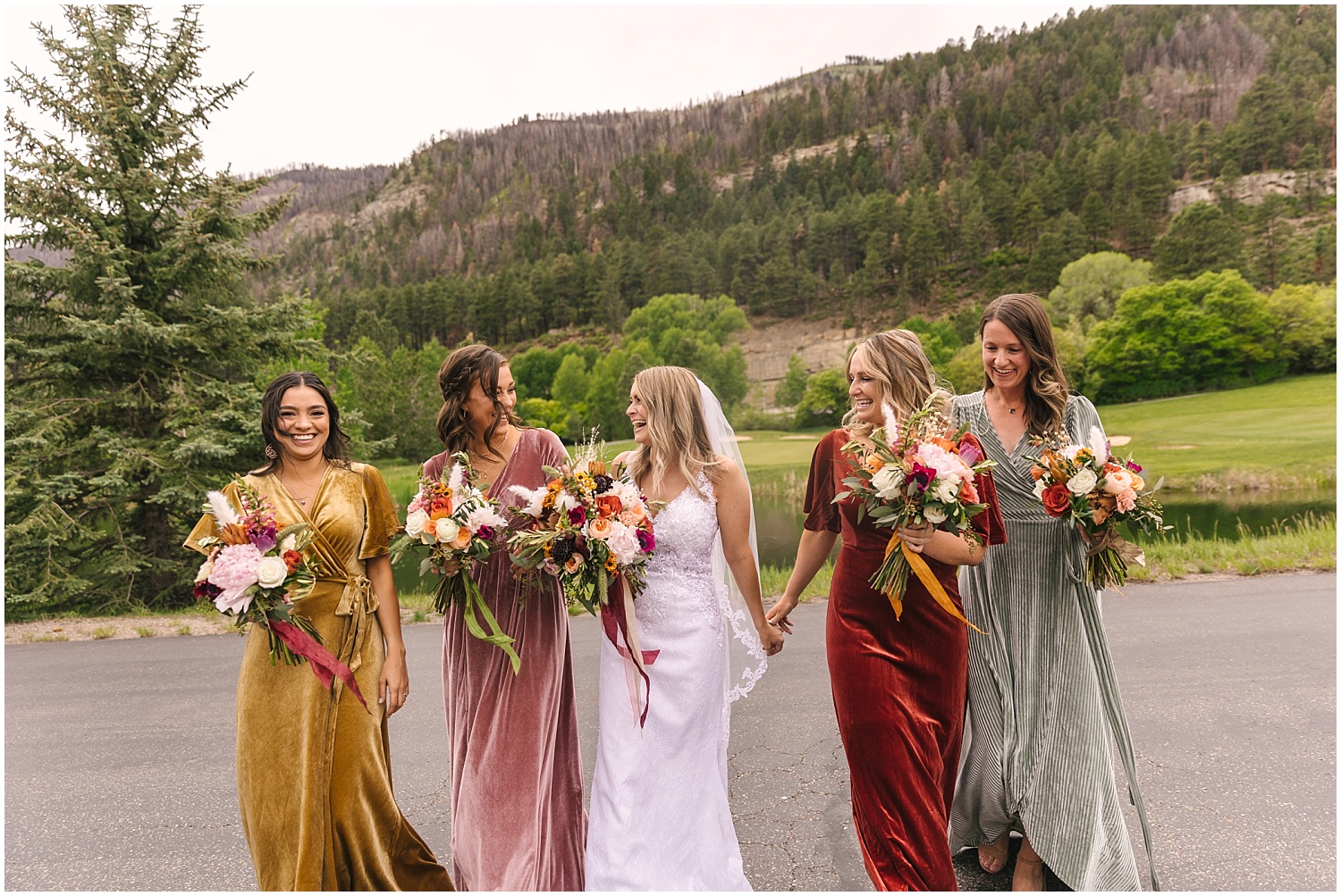 Bride walking with her bridesmaids in mismatched mustard, olive, burgundy, and pink velvet dresses at Glacier Club in Durango