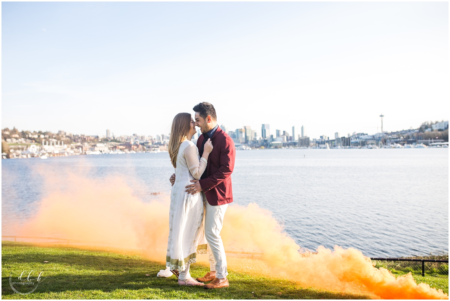 orange colored smoke bomb blowing around engaged Indian couple in front of Seattle skyline at Gas Works Park