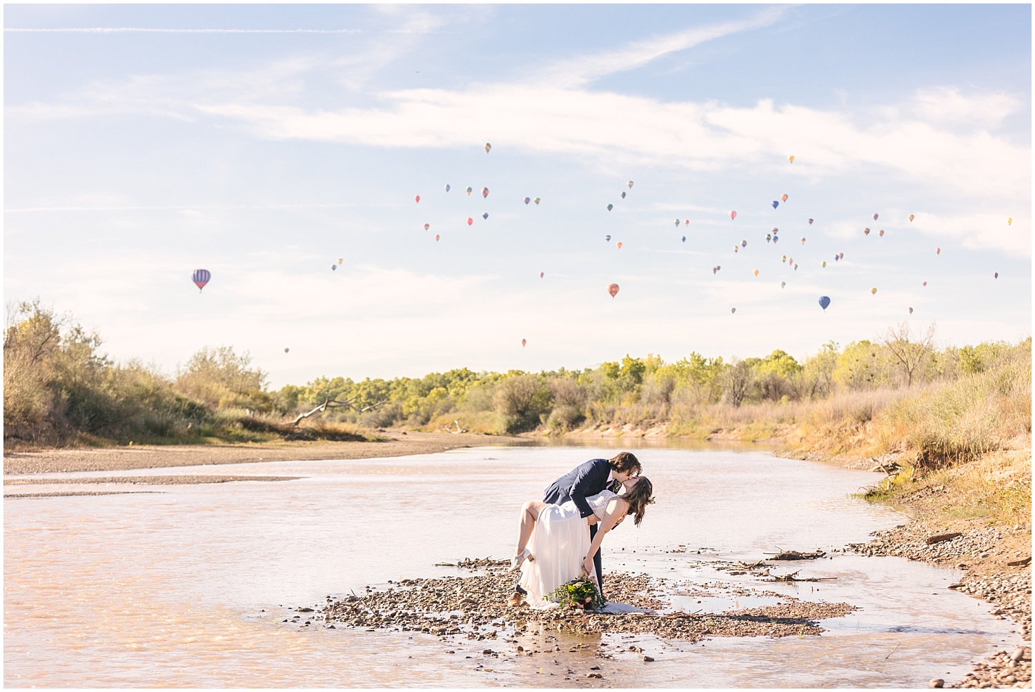 Bride and groom kissing in the Rio Grande river with hot air balloons in the background for their Albuquerque Balloon Fiesta elopement